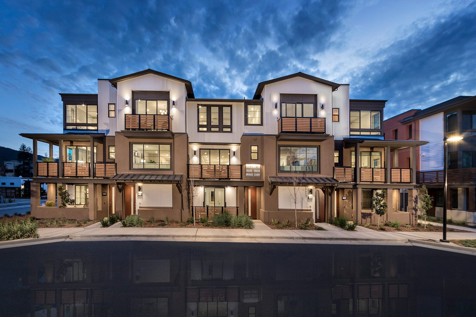 Modern row of stucco townhouses with illuminated windows at dusk. The clear blue sky and wet street create a peaceful, inviting atmosphere.