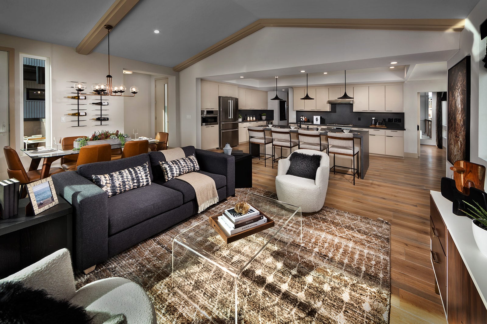 Stylish open-concept living room and kitchen with a cozy gray sectional, modern dining set, and elegant lighting. Warm tones and natural light create a welcoming vibe.