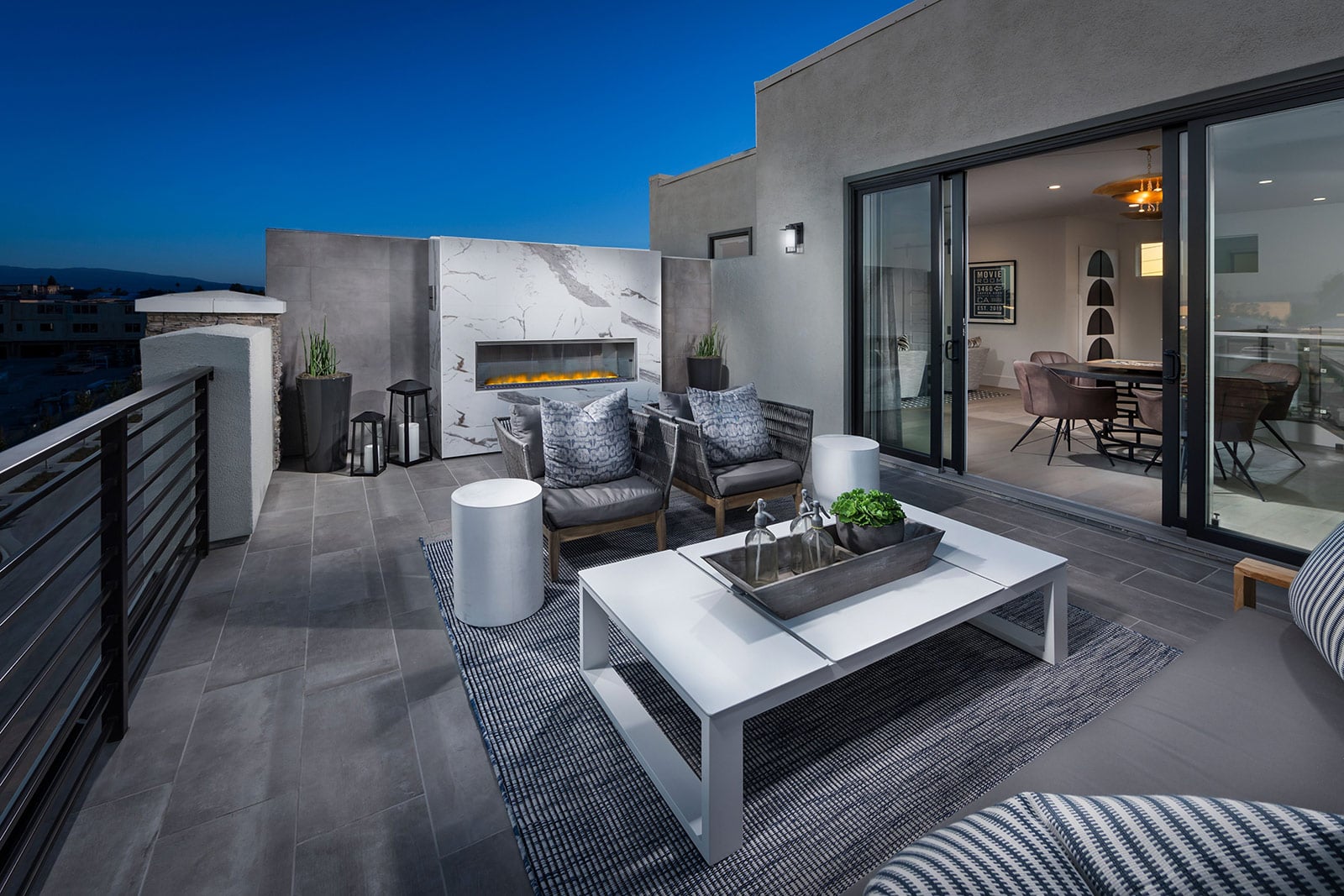 Outdoor living room with a modern fireplace, wall-mounted TV, cushioned seating, and patio chairs. A cozy backyard ambience with evening lighting.