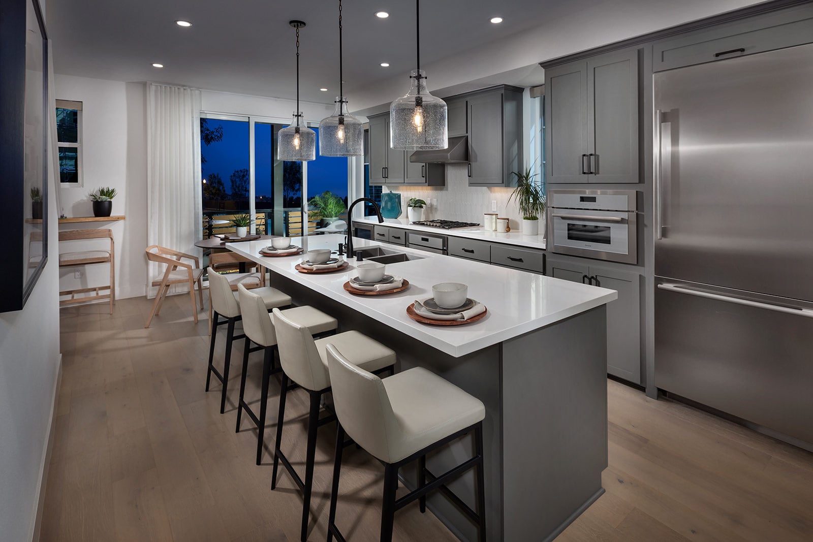 Modern kitchen and dining area with light wood flooring and gray cabinetry. A large table with black chairs sits on a rug. Pendant lights add warmth.